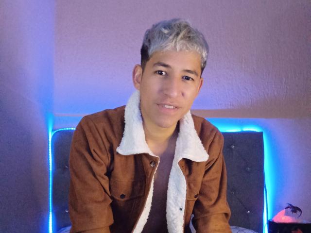 DantezSword, short-haired dude with blonde tips, wearing a brown suede jacket with white fur collar, sitting in front of blue neon lights, smiling slightly.