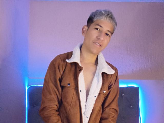 DantezSword with short gray hair, brown suede jacket, and white fur collar, slightly leaning forward, smiling subtly, blue LED lights in background.
