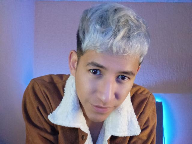 DantezSword with short, curly, blonde hair, brown eyes, and a cute smile, wearing a brown shirt with a white, fluffy collar, looking down with a slight tilt.