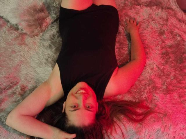 DayanHank lying on a fluffy grey rug, wearing a black one-piece, arms above head, smiling, red light on her, legs slightly apart, showing her ass.