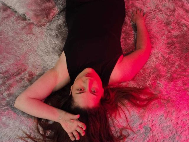 DayanHank lies on a fluffy grey rug, wearing a black sleeveless top, with red lighting on her face and arms, and her hand touching her head.
