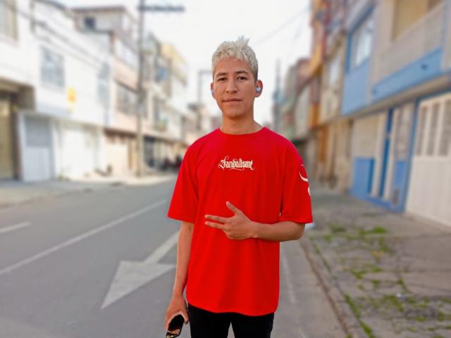 DantezSword with short blonde hair, red shirt, black pants, and a phone in hand, giving a thumbs-up. Standing on a street with blurred buildings in the background.