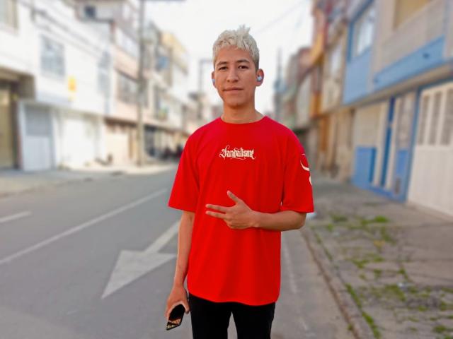 DantezSword with short, blonde hair stands in a red shirt, black pants, holding a black phone, right hand pointing, left ear with a white earbud, street in background.