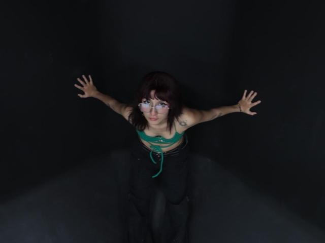 Ronnete with green bra, black pants, and tattoos, arms spread against dark walls, looking up with blue eye makeup, in dimly lit, narrow space.
