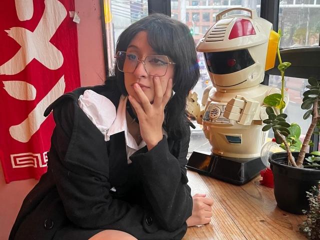 Ronnete with glasses, black hair, black jacket, white shirt, hand on cheek, sitting by robot, potted plant, wooden table, red and white flag, city view through window.
