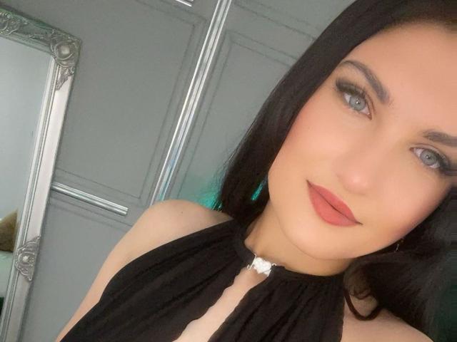 Elize Taylor tem pele clara, olhos azuis e cabelo preto. Ela está vestindo uma blusa preta com alças e um fecho branco, e tem batom vermelho nos lábios levemente entreabertos..