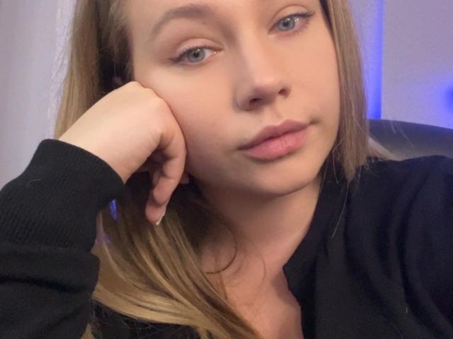 HollyCandyX with her hand on her cheek, blonde hair, blue eyes, pink lips, and black top, looking slightly to the side with a relaxed expression.