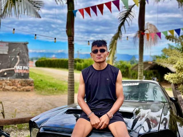 HotBrunoX with short black hair, wearing a sleeveless black shirt and black shorts, sitting on a black car, sunglasses on, muscular, tan skin, palm trees, colorful flags in background.