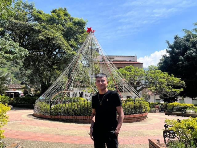 HotBrunoX standing in front of a wire dome with a red star on top, wearing a black shirt, hands in pockets, short dark hair, blue sky, green trees.