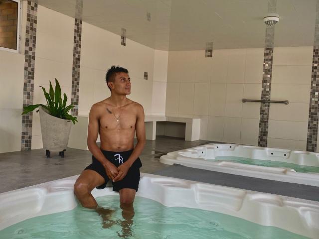 HotBrunoX, shirtless, in a jacuzzi, wearing black shorts, hands in front, looking up, tattoo on chest, short dark hair, medium brown skin, small chest, potted plant in background.