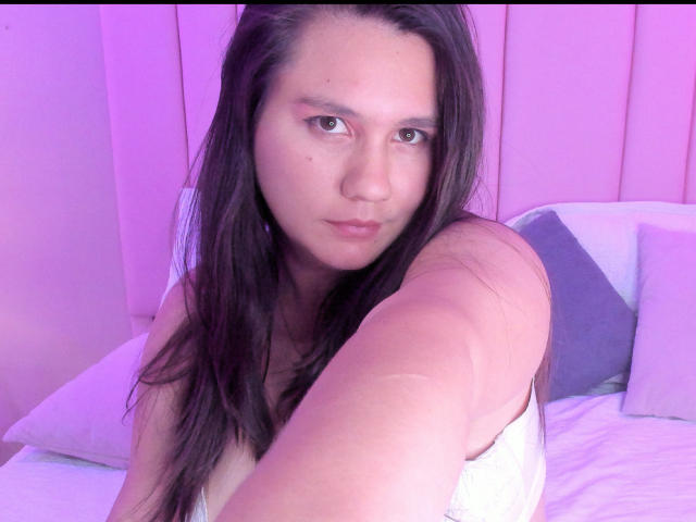 AvienLume mit langen, braunen Haaren, rosafarbener Haut und dezentem Make-up, sitzt auf einem Bett mit weißen Laken, trägt ein weißes Oberteil und macht ein Selfie, wobei sie einen neutralen Gesichtsausdruck zeigt..