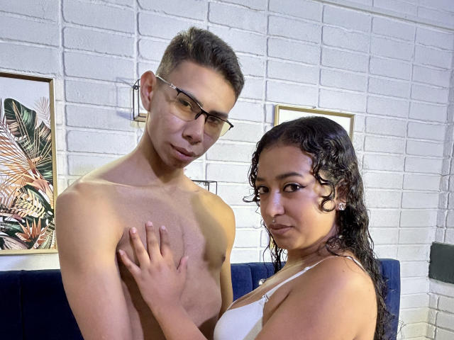 IsaAngel69, con cabello negro rizado, un sostén blanco y piel morena, tocando a IsaAngel69, que está sin camisa, con gafas y cabello castaño corto, frente a una pared de ladrillos blancos..