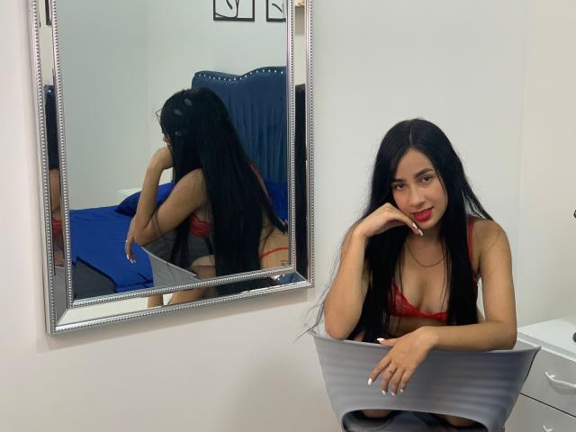 KityLoven, avec de longs cheveux noirs, en lingerie rouge, assise, souriante, la main sur le menton, regardant dans un miroir. Le reflet montre ses fesses. Mur blanc, chaise bleue, cadre de miroir argenté, bol gris..