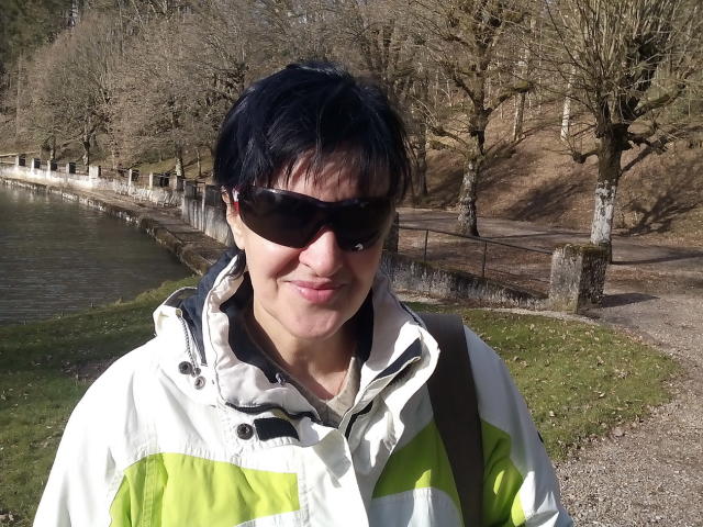 ReniaHot with short black hair, wearing sunglasses and a white-green jacket, has a slight smile and is standing by a waterway. Her backpack is on her right.