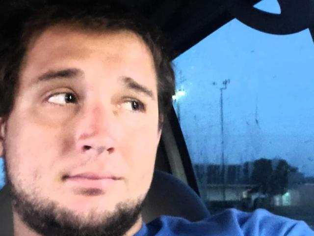 DoyleRobert69 has a slightly confused, thoughtful expression with a short beard, brown curly hair, and is wearing a blue shirt, looking out of a car window.