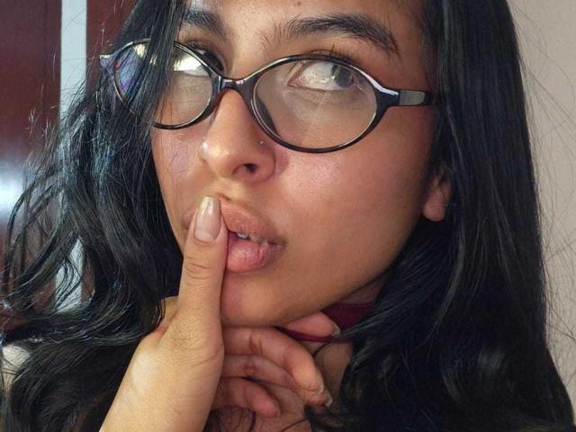 AdaWalton with dark hair, glasses, and nose ring, pouting with finger on lips, has light brown skin, and is looking up. Her hand is near her chin.