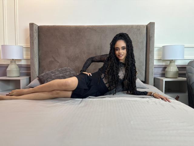 TerryGreen, with long curly black hair, lies on a bed, wearing a sheer black top and black shorts, showing her bare legs and a subtle smile.