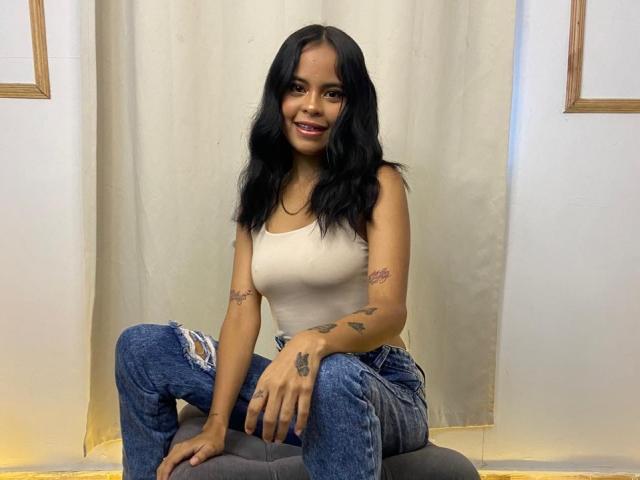 BellaLena with black wavy hair, tan tank top, and ripped blue jeans, sitting on a gray cushion, smiling with small tattoos on her arms and hands.