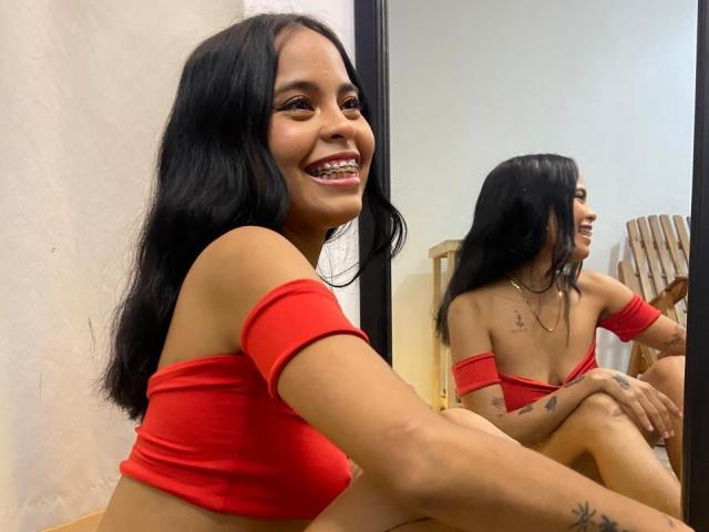 BellaLena with long black hair, smiling with braces, wearing a red off-shoulder top, reflecting in a mirror, sitting with her knee up, showing tattoos on her arm.