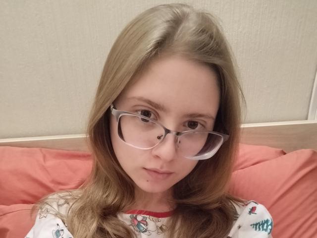 DarkLilith69, com cabelos loiros longos, usando óculos e uma camisa branca com padrões coloridos, tem uma pequena pinta acima dos lábios levemente entreabertos, sentada em almofadas rosa..