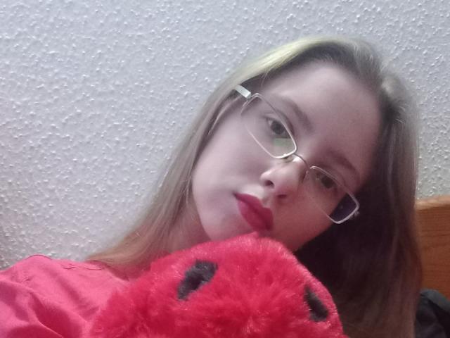 DarkLilith69, avec des lunettes, des cheveux blonds et du rouge à lèvres, tenant un jouet en peluche rouge et duveteux contre sa poitrine, portant une chemise rose, regardant la caméra..