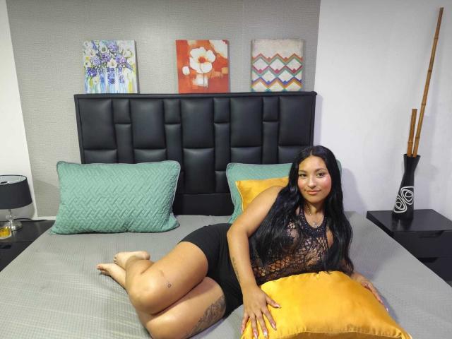 CelesteTower, com longos cabelos pretos, pele bronzeada e tatuagem na coxa, deitada na cama, vestindo uma blusa preta de renda e shorts pretos, segurando um travesseiro amarelo, com travesseiros verdes e cinzas atrás..
