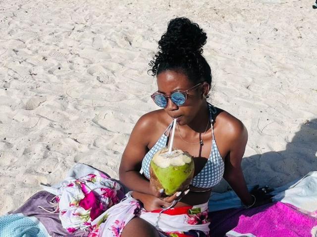 SexyValentinaLove with dark curly hair in a bun, wearing a checkered bikini top and floral skirt, sipping from a green coconut on a sandy beach, wearing round sunglasses.