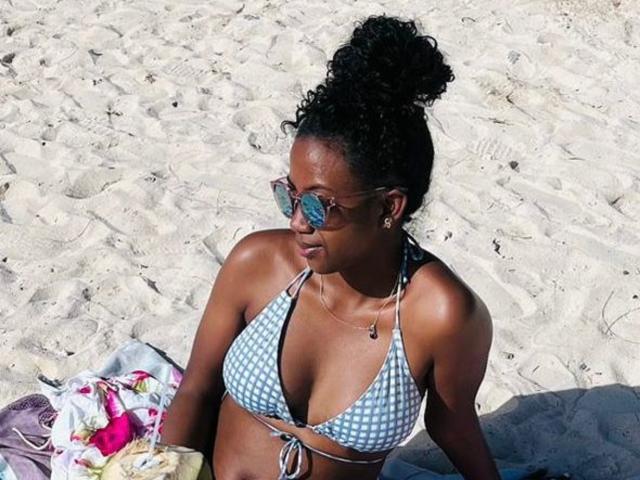 SexyValentinaLove has dark skin, curly black hair in a bun, wears a blue checkered bikini top, and reflective sunglasses, sitting on a sandy beach with a towel.