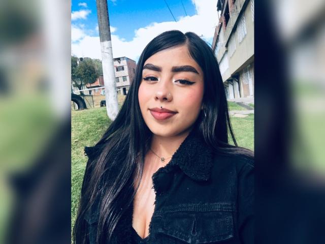 Julie Addams, avec ses longs cheveux noirs, ses sourcils audacieux et son rouge à lèvres rose, portant une veste en jean noir, se tient à l'extérieur avec un arrière-plan flou, et a l'air confiante et séduisante..
