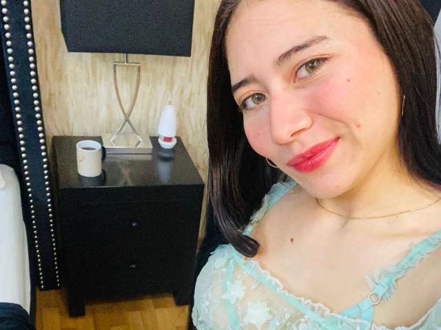 LexyJones, con le labbra rosa e gli occhi marroni, indossa una camicetta trasparente di pizzo blu che mette in mostra il seno. Ha una piccola lentiggine sulla guancia destra e una collana d'oro..