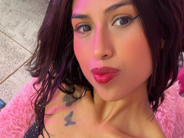 MyaaTyler with pink lipstick, dark eyeliner, and wavy brown hair, showing off butterfly tattoos on her chest. She's on a pink fluffy rug with a tile floor.