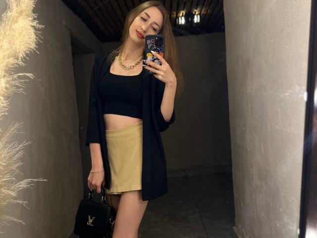QueenKiss with long blonde hair, black crop top, gold skirt, black cardigan, gold necklace, holding black handbag, taking mirror selfie, smiling, holding phone with star pattern case.