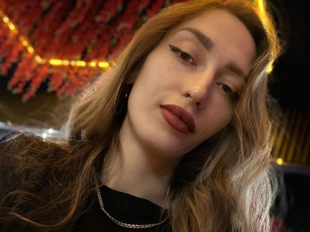 QueenKiss has wavy blonde hair, thick eyebrows, black eyeliner, and red lipstick. She wears a black top and a silver chain, with a slightly bored expression.