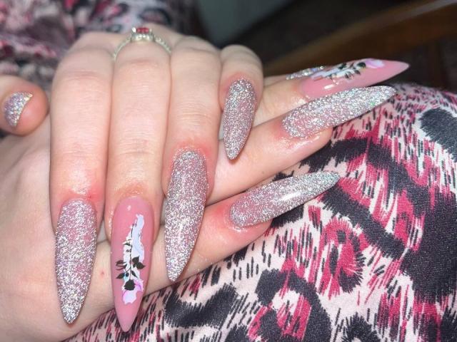 LaPulia's hands with long, glittery pink nails, featuring a black and white floral design on the ring nail. She's wearing a patterned pink and black garment.