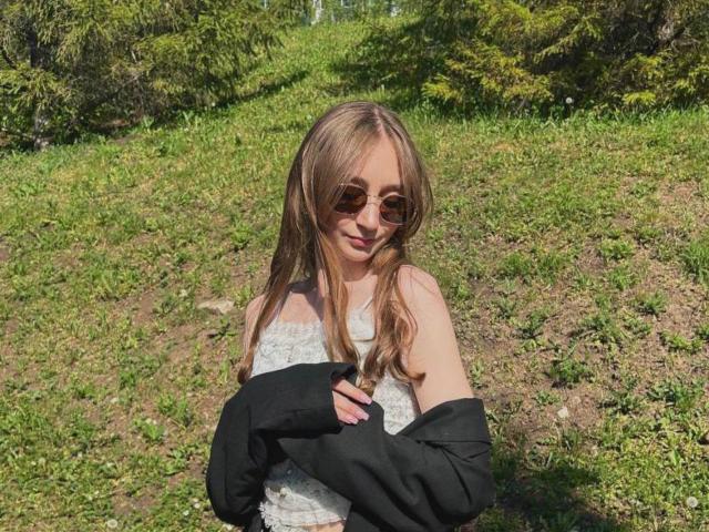 CynthiaWhite with long blonde hair, wearing sunglasses, a white lace dress, and a black jacket, standing in a grassy area, smiling slightly. Her tits are small and she's cute.
