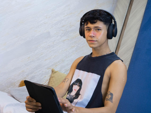 DanteEvans, with curly black hair and headphones, wears a black sleeveless shirt with a portrait, holding a tablet, has tattoos on both arms, sitting on a bed.
