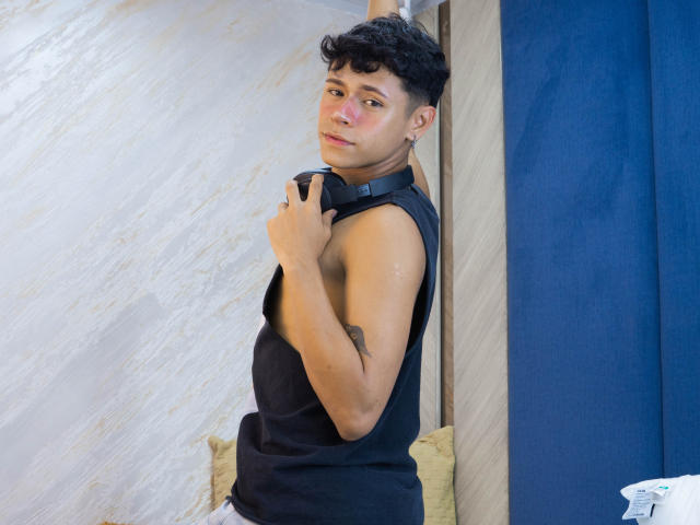 DanteEvans with short curly black hair, light brown skin, and a black sleeveless shirt, holding a black strap against a blue curtain. His right arm has a small tattoo.
