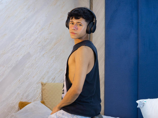 DanteEvans, a young dude with curly black hair, wearing a black sleeveless shirt and headphones, sits on a bed, looking over his shoulder with a slight smirk.