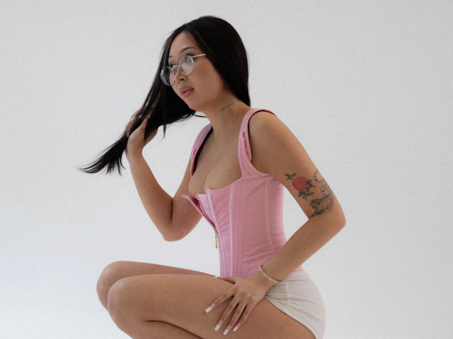NaomiAsian with black hair, glasses, pink corset, white shorts, tattooed arm, holding hair, squatting, showing small tits, looking to the side.