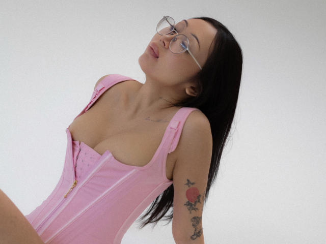 NaomiAsian with glasses, pink corset, and long black hair, head tilted back, showing small tits and tattoo on right arm, against white background.