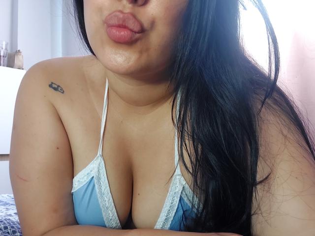 LauraRichis with pouty lips, black hair, light skin, blue lace-trimmed bra, showing large tits, tattoo on right shoulder, light pink bedroom background.