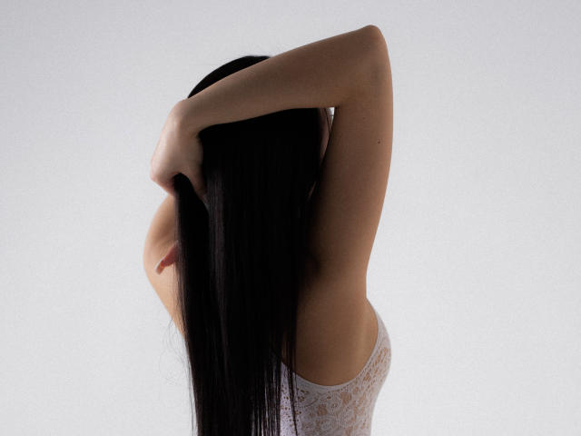 NaomiAsian with long black hair, arms raised, hands in hair, wearing a white lace top, showing her bare back and small, perky tits. White background.