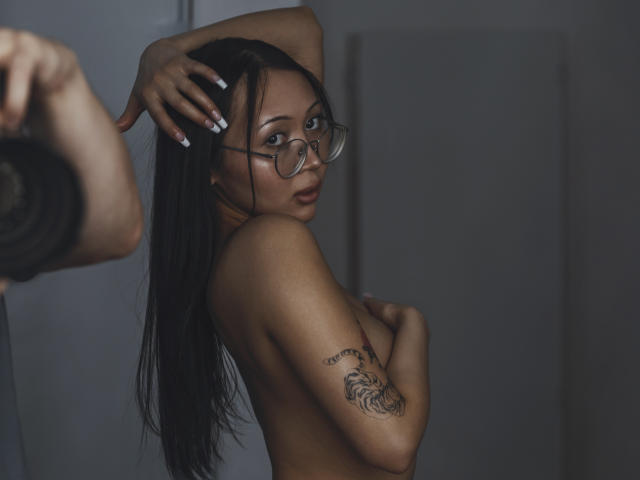 NaomiAsian with long black hair, glasses, and a tattoo on her upper arm, topless, covering her tits with one arm, looking over her shoulder. Dark, moody photograph.