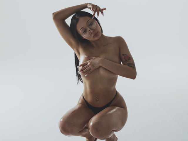 NaomiAsian, topless, wearing black panties, kneeling, covering small tits with hand, arm raised, glasses, tattoo on right arm, belly button piercing, wet skin, long black hair.
