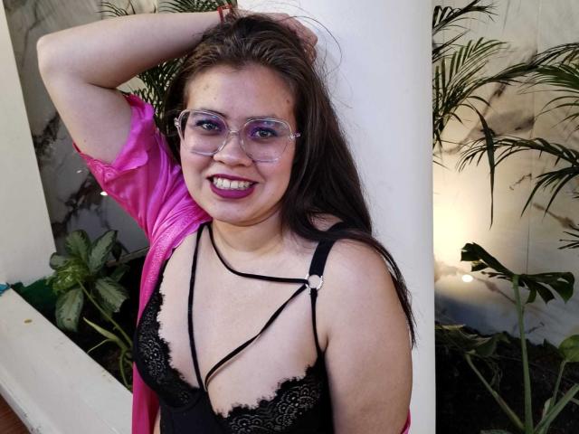 Isabella Reid con gli occhiali, una camicia rosa, un reggiseno di pizzo nero, i seni scoperti, rossetto viola, che sorride, con un braccio alzato, capelli castani, in piedi accanto a un pilastro bianco, piante verdi sullo sfondo..