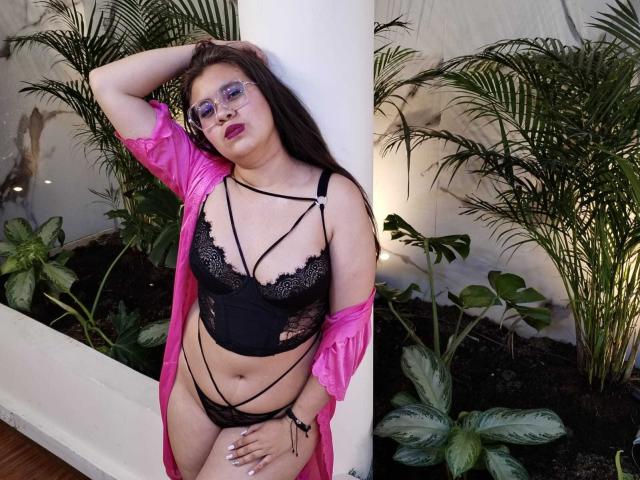 Isabella Reid, avec des cheveux foncés, des lunettes, une robe rose ouverte, de la lingerie noire en dentelle, des culottes noires à fines bretelles, une main dans les cheveux, du rouge à lèvres rose, debout, des plantes vertes, un mur blanc, un sol en bois..