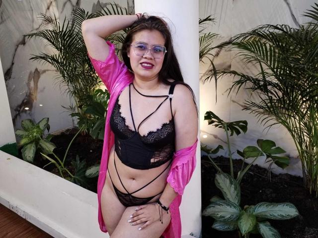IsabellaReid with glasses, pink robe, black lace lingerie, strappy, showing her tits and pussy, arm up, hand on white column, green plants behind. Smiling, dark hair, fair skin.