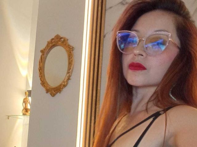 AngelinaBlanche hat rotes Haar, trägt klare Brillen und roten Lippenstift. Sie trägt ein schwarzes Top mit dünnen Trägern, das ihre Brüste zeigt. Im Hintergrund befindet sich ein Spiegel mit goldener Rahmen..