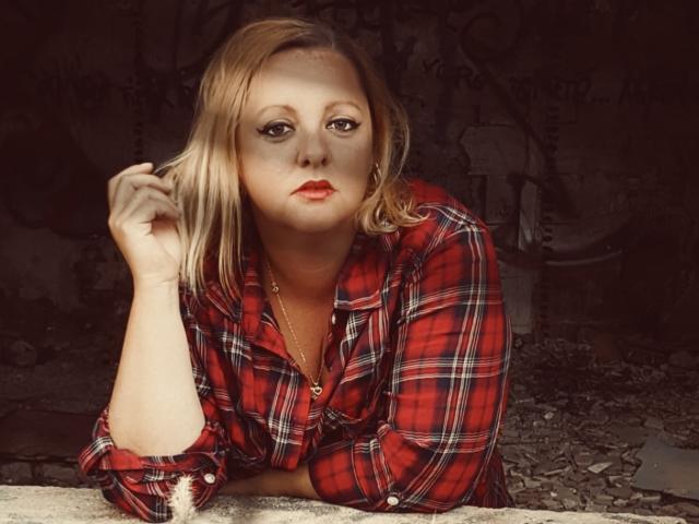 CplCokins with blonde hair, red plaid shirt, and red lipstick, leaning on a broken wall, holding her hair with a serious expression, wearing a gold necklace.