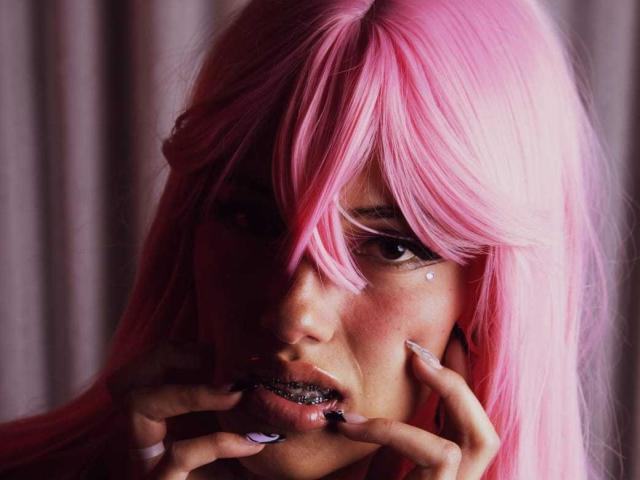 SharlotteX, com cabelo rosa, batom escuro e glitter nos lábios, dedos perto da boca, com uma expressão sedutora, com um olho ligeiramente escondido pela franja. A maquiagem dela é escura e dramática..
